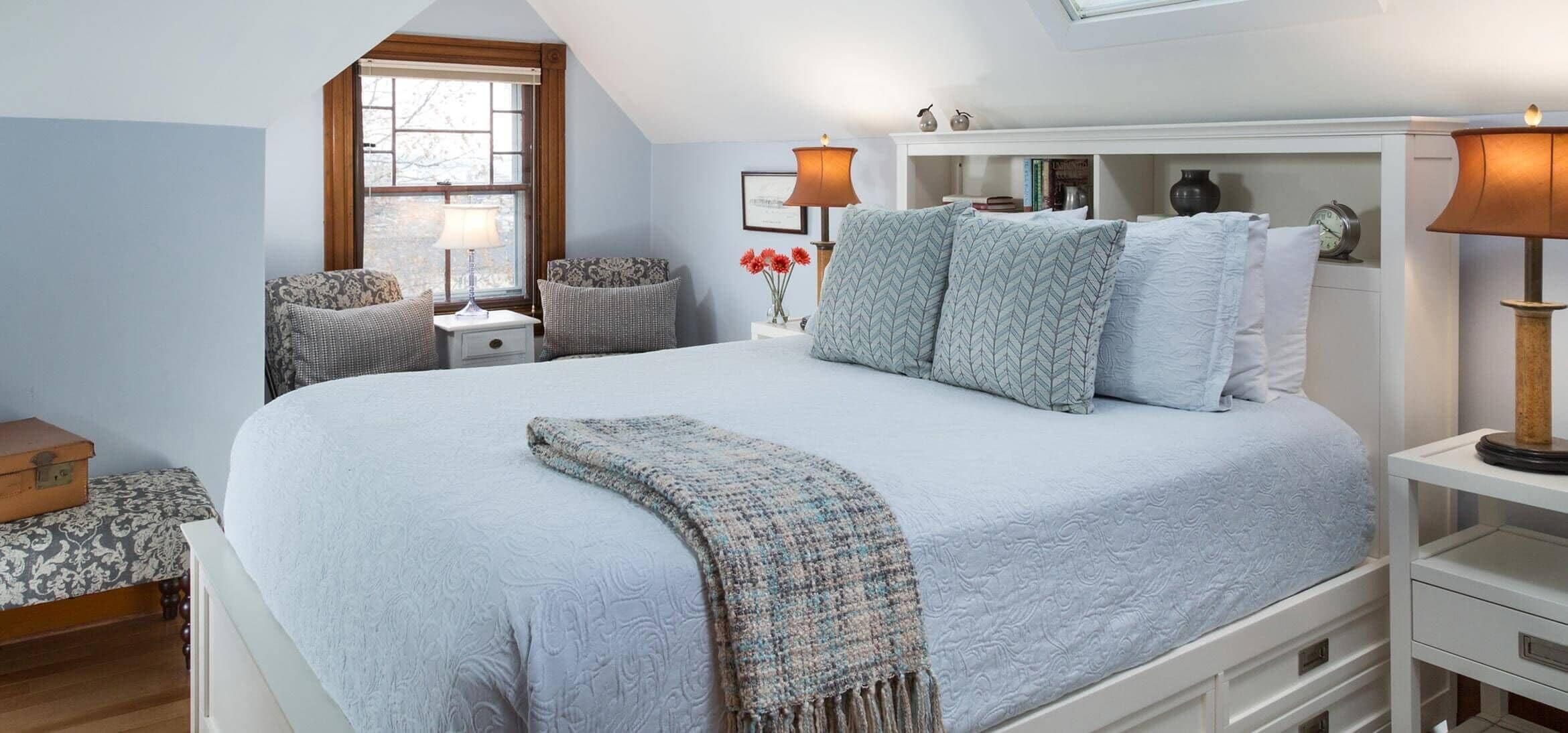 A bedroom with a large bed, a nightstand with a lamp, a chest of drawers, a window, and a bookshelf.