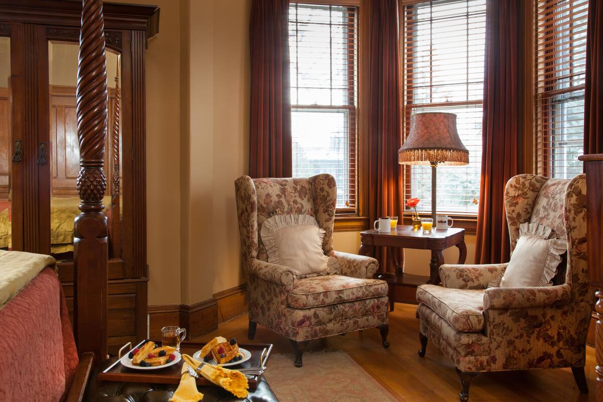 A hotel room with a large bed, two floral armchairs, a small table with a lamp, and a tray with breakfast items.