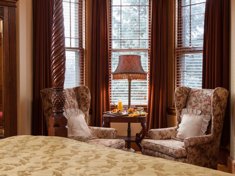 A room with two upholstered chairs, a wooden table with a lamp, and a bed with a patterned bedspread.