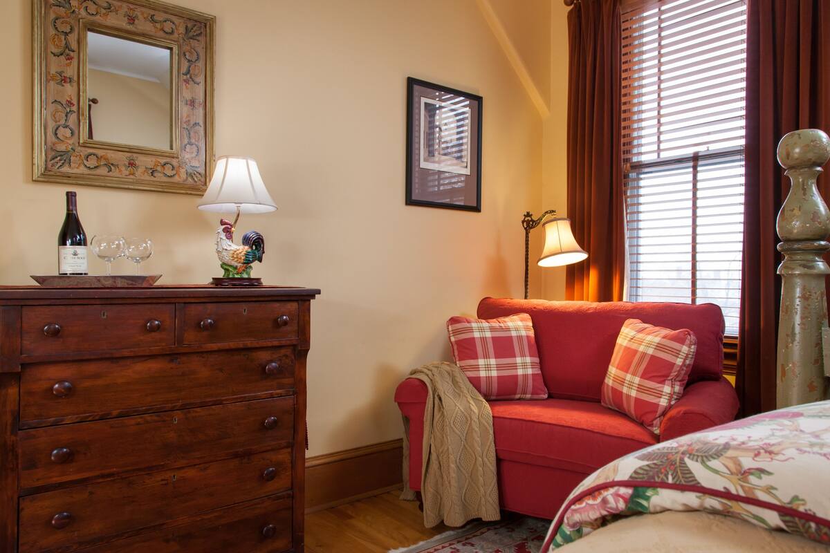 A cozy room with a wooden dresser, a red sofa with plaid cushions, a framed mirror, and a window with blinds.