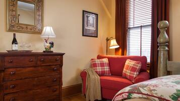 A cozy room with a wooden dresser, a red sofa with plaid cushions, a framed mirror, and a window with blinds.