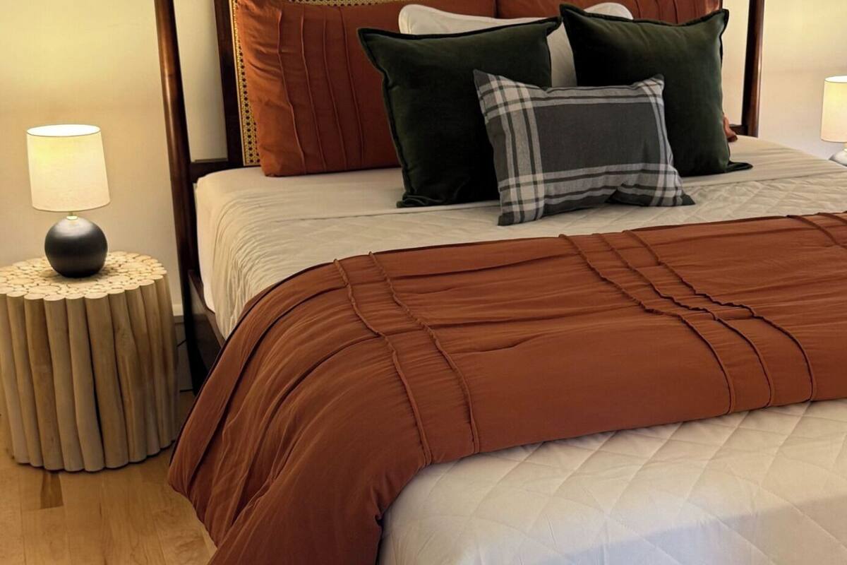 A neatly made bed with a brown blanket, a pillow, and a bedside lamp on a round wooden nightstand.