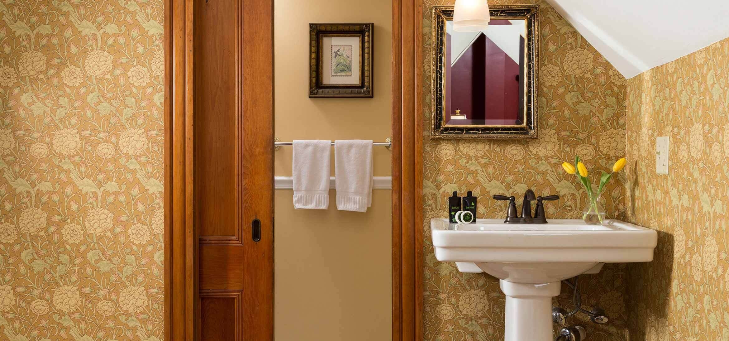 A bathroom with floral wallpaper, a wooden door, a white sink with toiletries, and a mirror with a decorative frame.