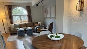 A modern living room with a round wooden table, grey upholstered chairs, a beige sofa, and a large window with curtains. - Lang House On Main Street (Burlington)