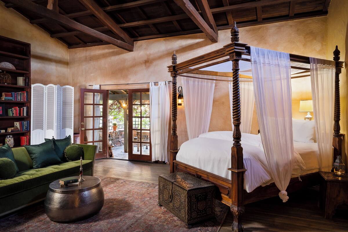A four-poster bed with white linens, a green sofa, a wooden coffee table, and a bookshelf with books.