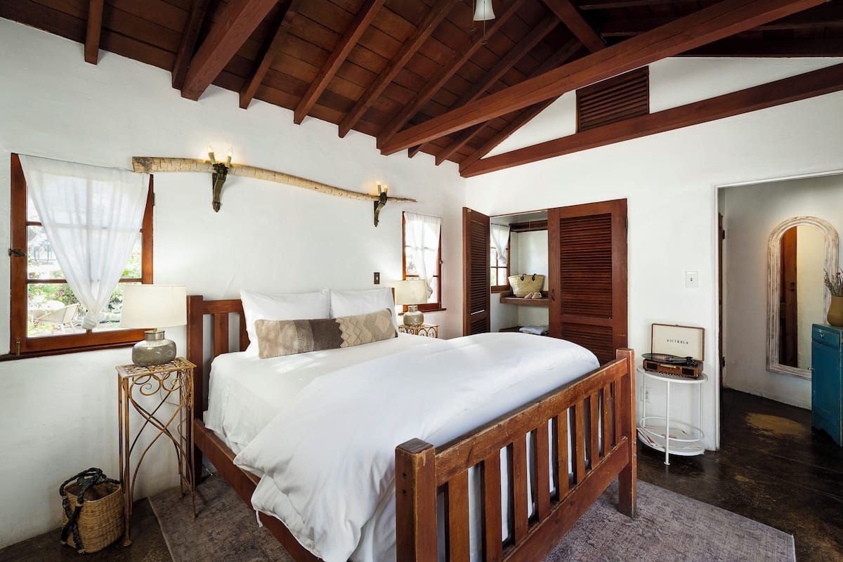 A bedroom with a wooden bed, a nightstand, a lamp, a window with sheer curtains, and a wooden ceiling.