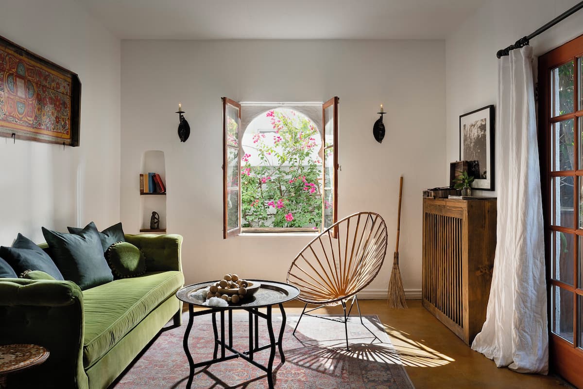 A room with a green sofa, a round table, a wooden chair, a window with a view of pink flowers, and a cabinet with a curtain.
