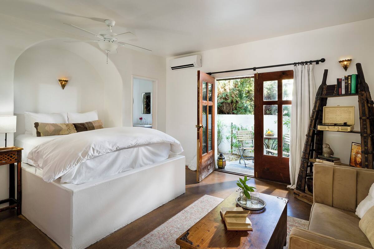 A bedroom with a large bed, a ceiling fan, an air conditioning unit, a wooden side table, a bedside lamp, a small table with books and a plant, and a view of the outdoors through a glass door.