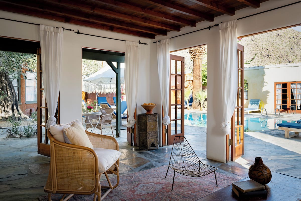 A room with wicker furniture, a small table, and a chair. There is a window with curtains, a door leading outside, and a view of a pool.