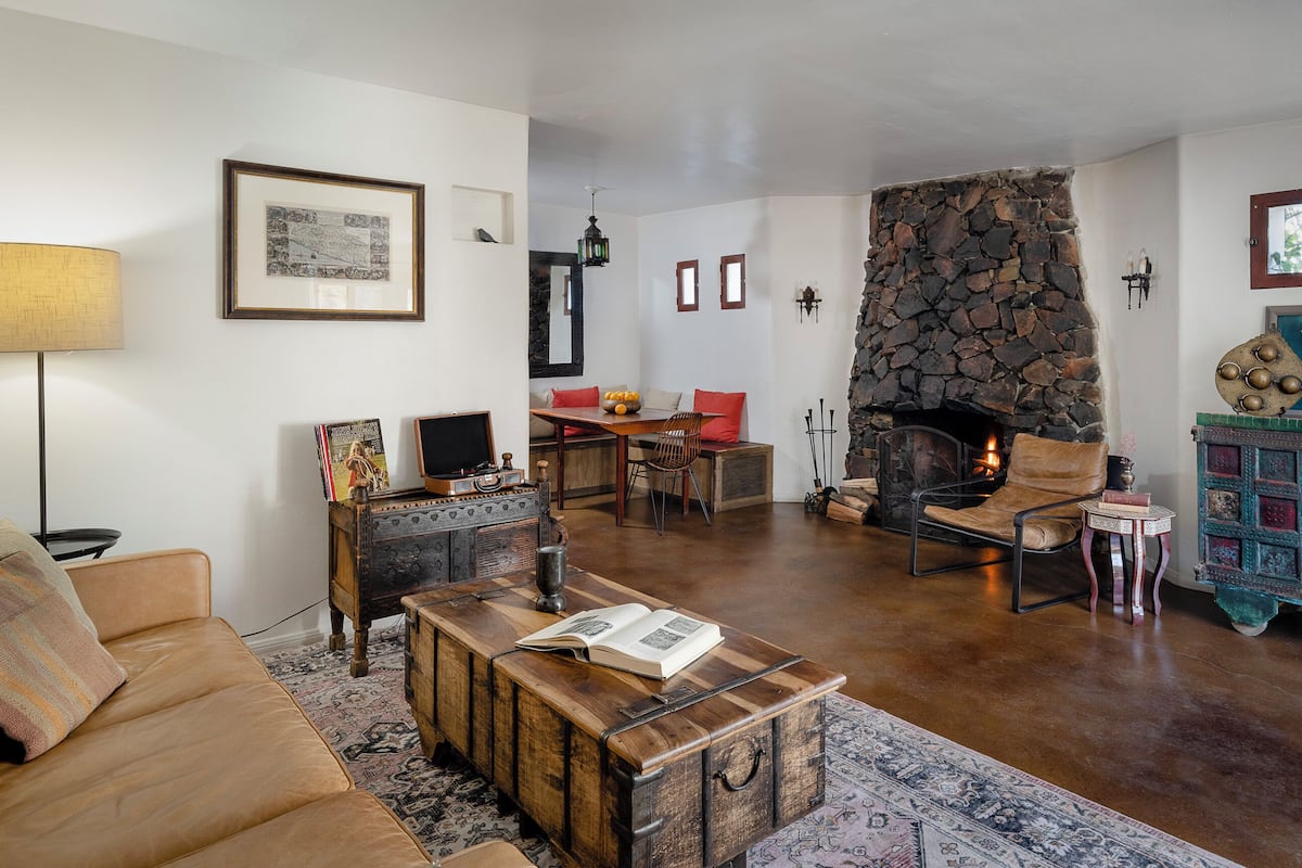A cozy living room with a stone fireplace, a wooden coffee table with books, a leather sofa, and a patterned rug.