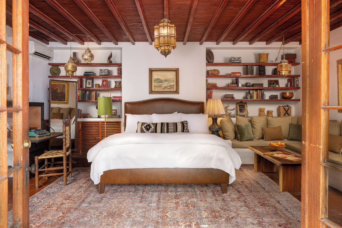 A cozy bedroom with a large bed, wooden headboard, and a patterned rug. There are shelves with books and decorative items, and a wooden bench with a basket of fruit.