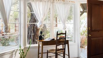 A room with a wooden chair, a table with a book, and a potted plant by a window with white curtains.