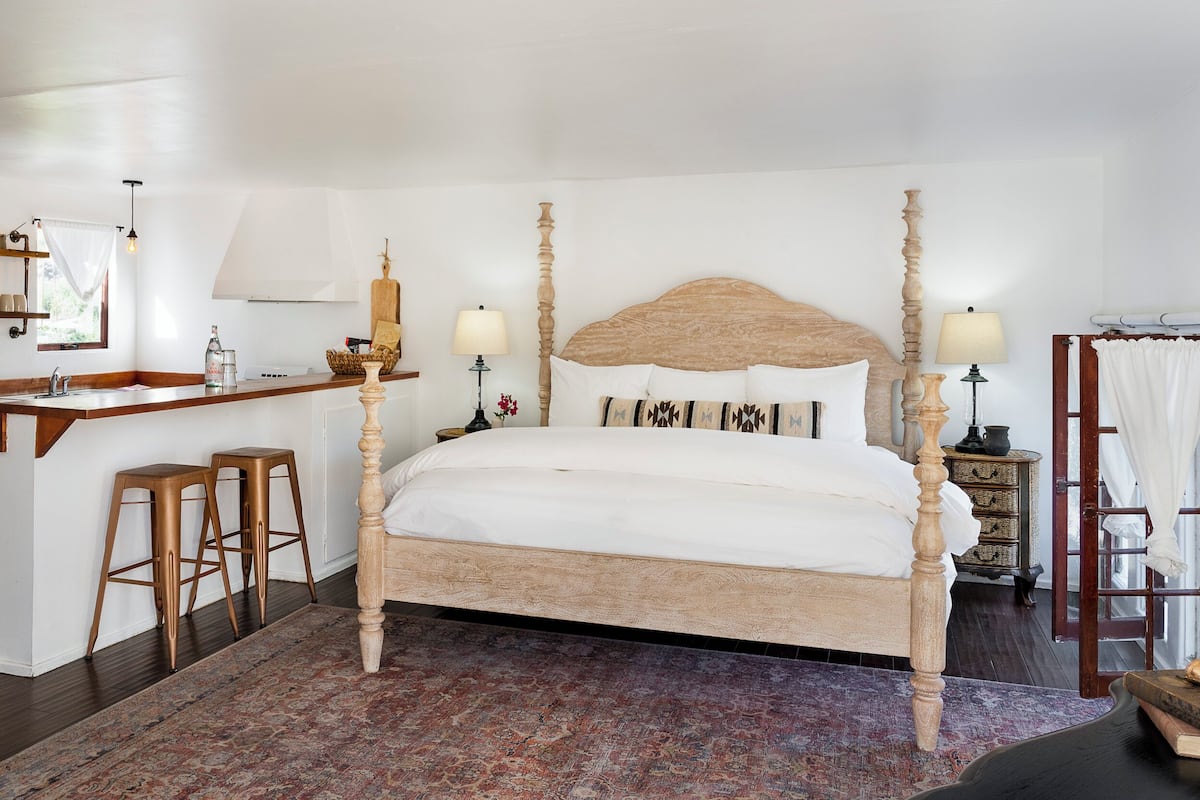 A spacious bedroom with a four-poster bed, a wooden desk with stools, a small kitchen area, and a large window.
