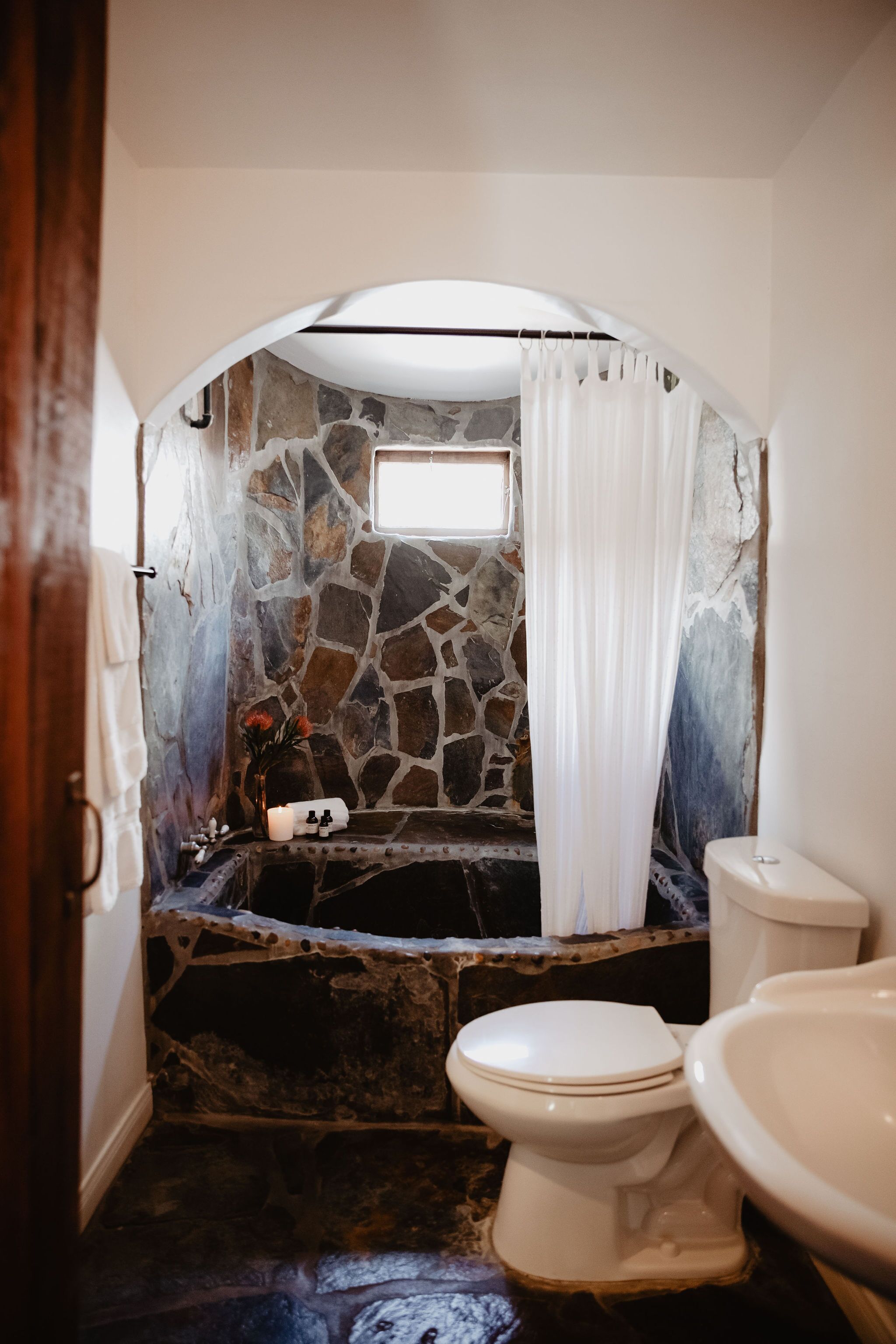 A bathroom with a stone wall, a bathtub, a toilet, and a shower curtain.