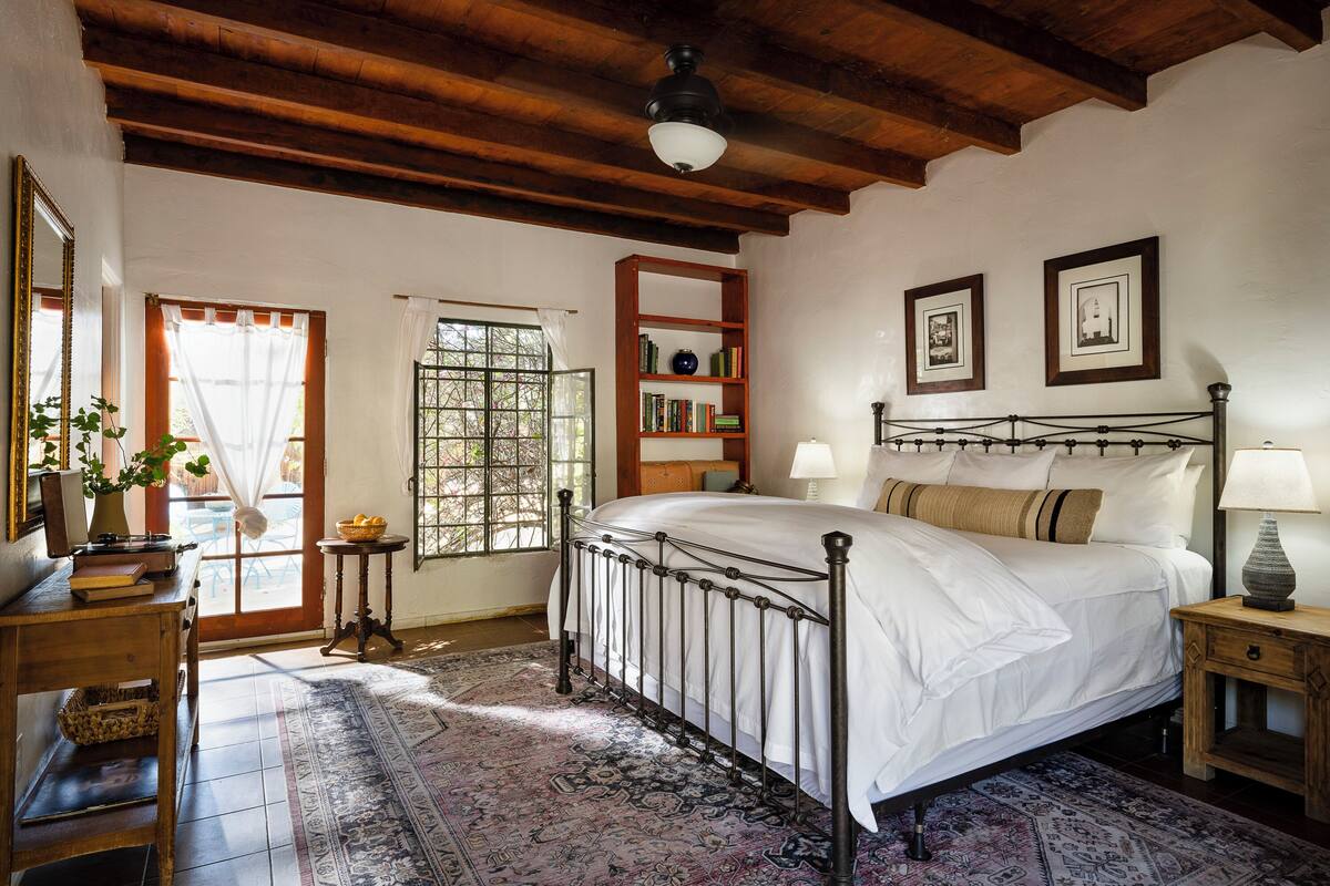 A bedroom with a metal bed frame, a wooden headboard, a bedside table with a lamp, a bookshelf, and a large window with curtains.