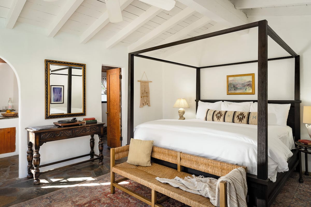 A four-poster bed with white linens, a wooden bench, a console table with a mirror, and a patterned rug.