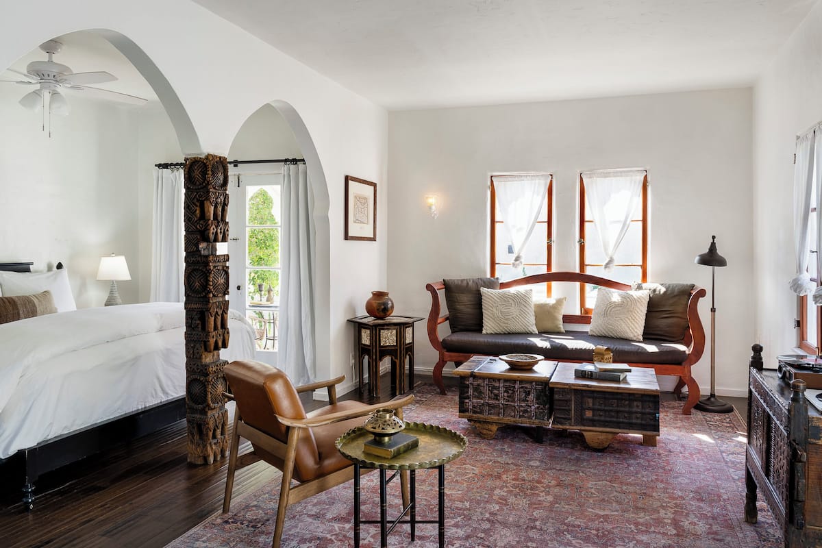 A room with a bed, a sofa, a coffee table, and a small table with a lamp. There are windows with curtains and a ceiling fan.