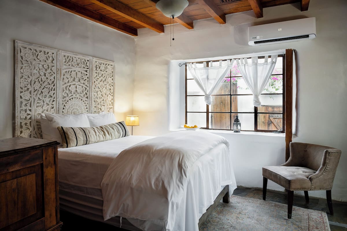 A bedroom with a bed, a chair, a wooden dresser, a window with curtains, and an air conditioning unit.