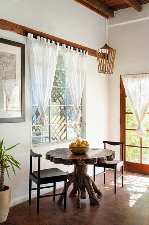 A rustic dining area with a wooden table and chairs, a hanging light fixture, and a window with white curtains.