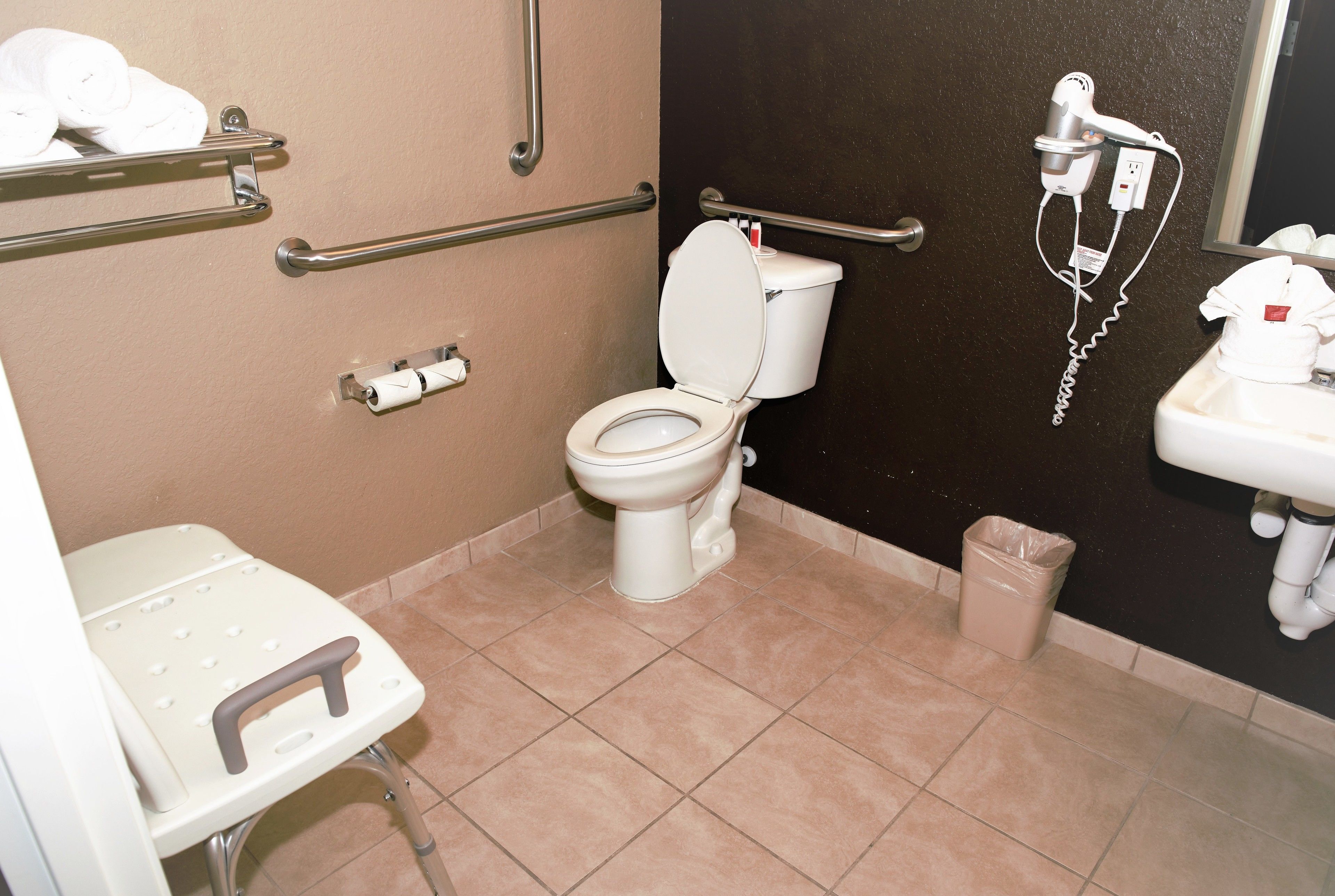 A bathroom with a toilet, handrails, a step stool, a trash can, and a hairdryer on the wall.