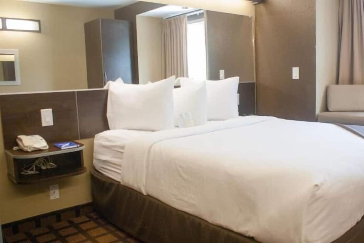 A hotel room with a large bed, a bedside table with a phone, a mirror, and a window with curtains.