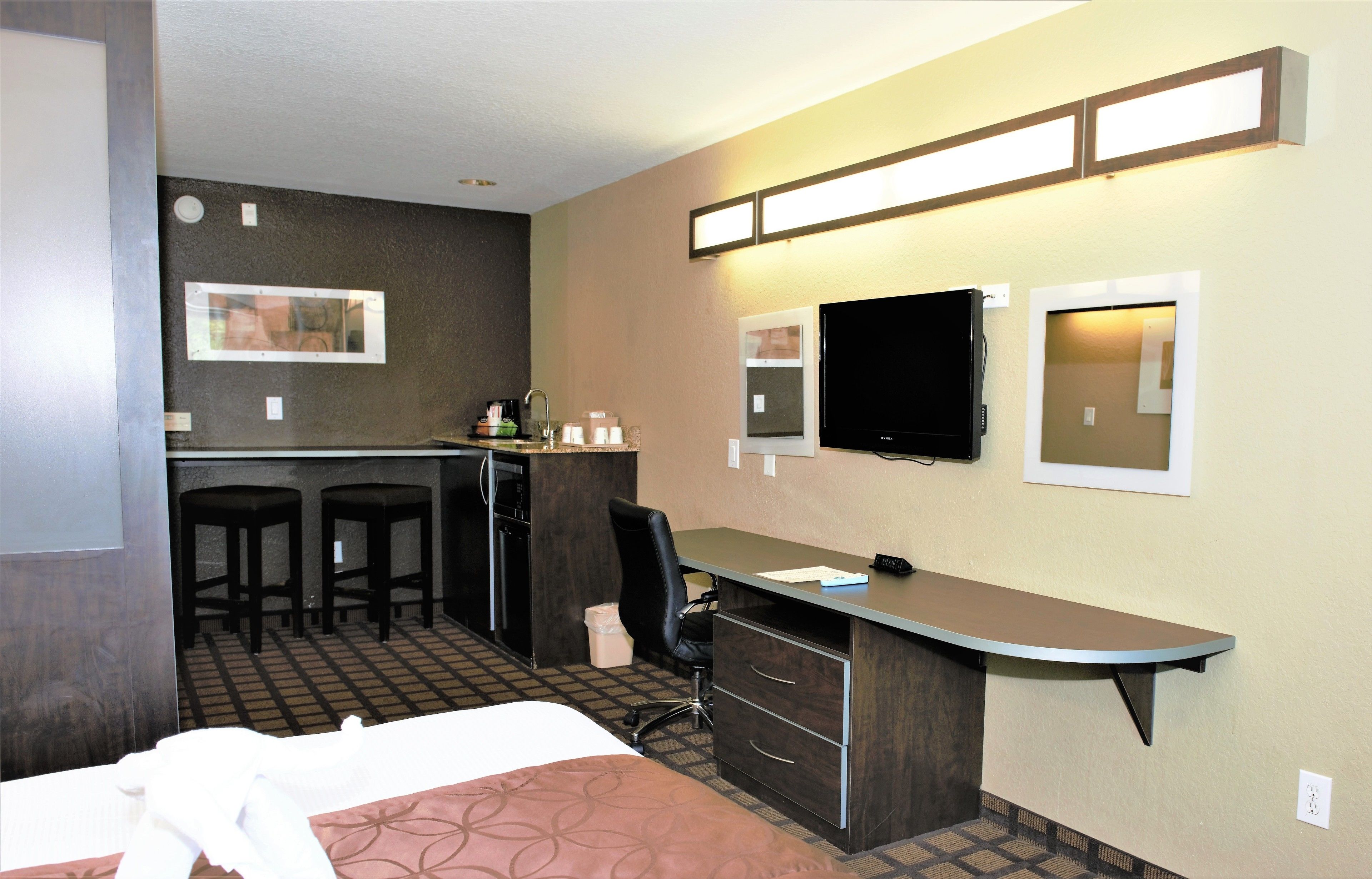 A hotel room with a bed, desk, two bar stools, a television, and a small kitchenette.
