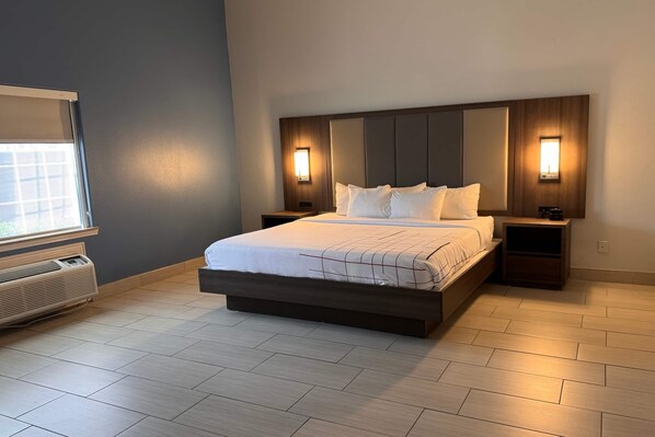 A modern hotel room with a large bed, built-in headboard, bedside tables, and a window with blinds.