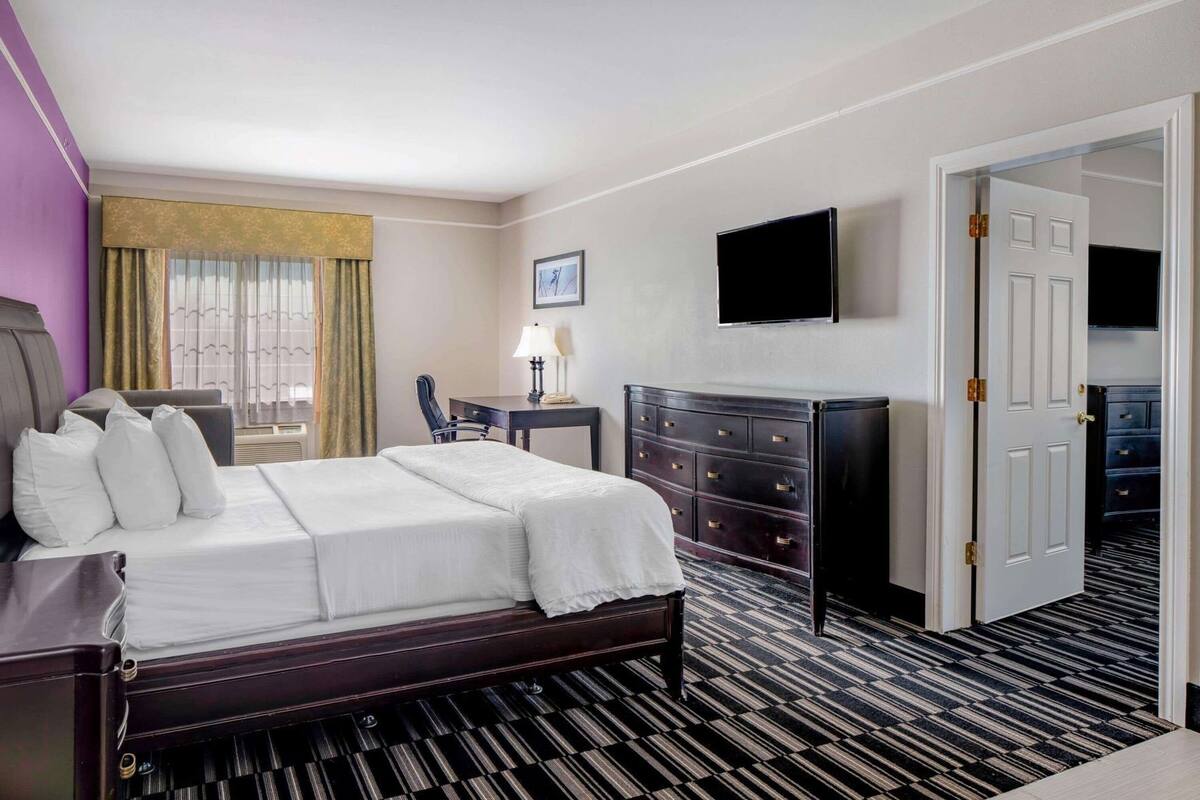 A hotel room with a bed, a desk, a dresser, a TV, and a door leading to another room.
