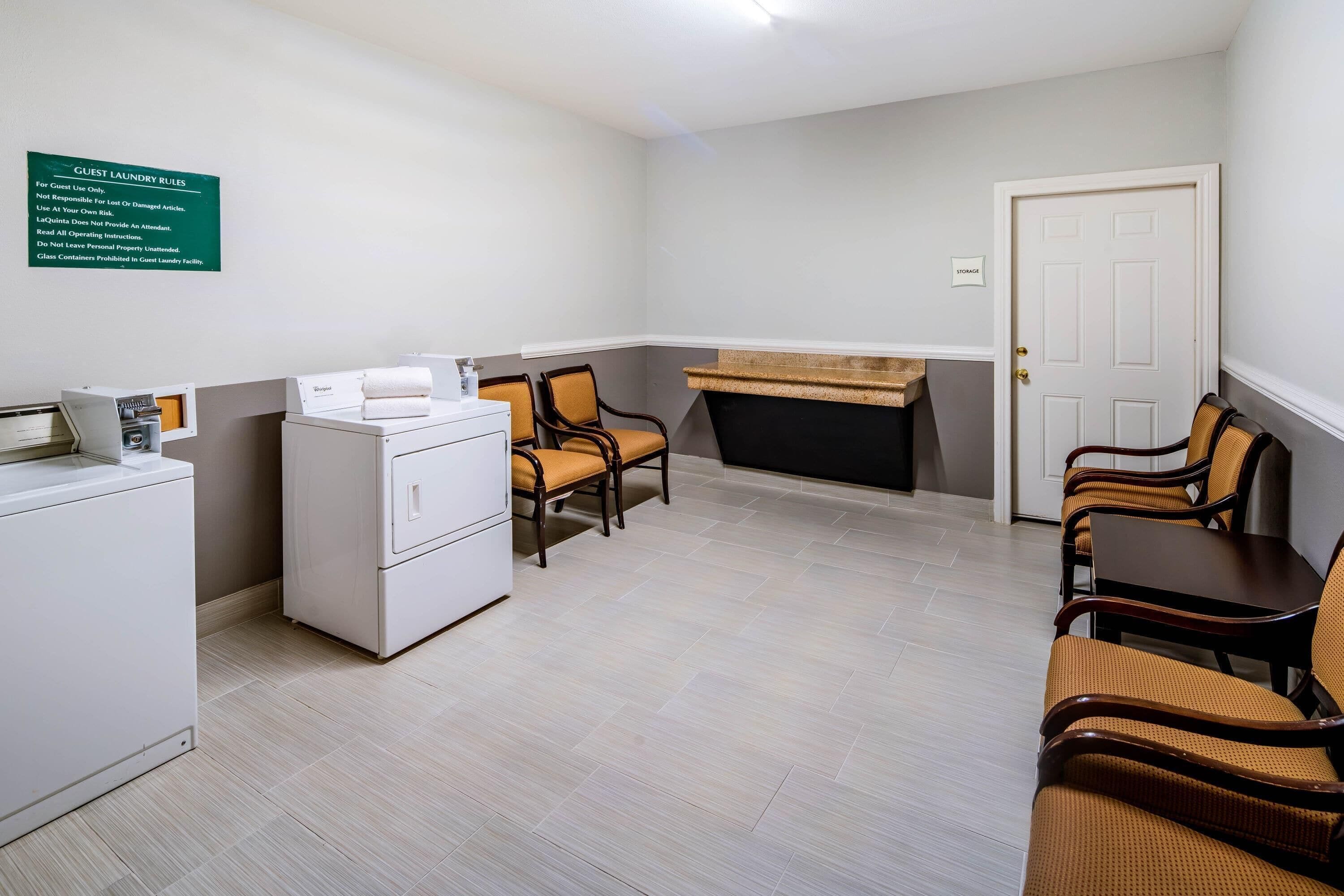 A guest laundry area with a washing machine, dryers, and a sign with laundry rules. There are chairs and a table in the room.