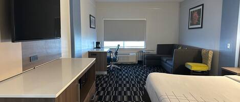 Una habitación de hotel con cama, escritorio, silla, televisor y ventana con persianas.