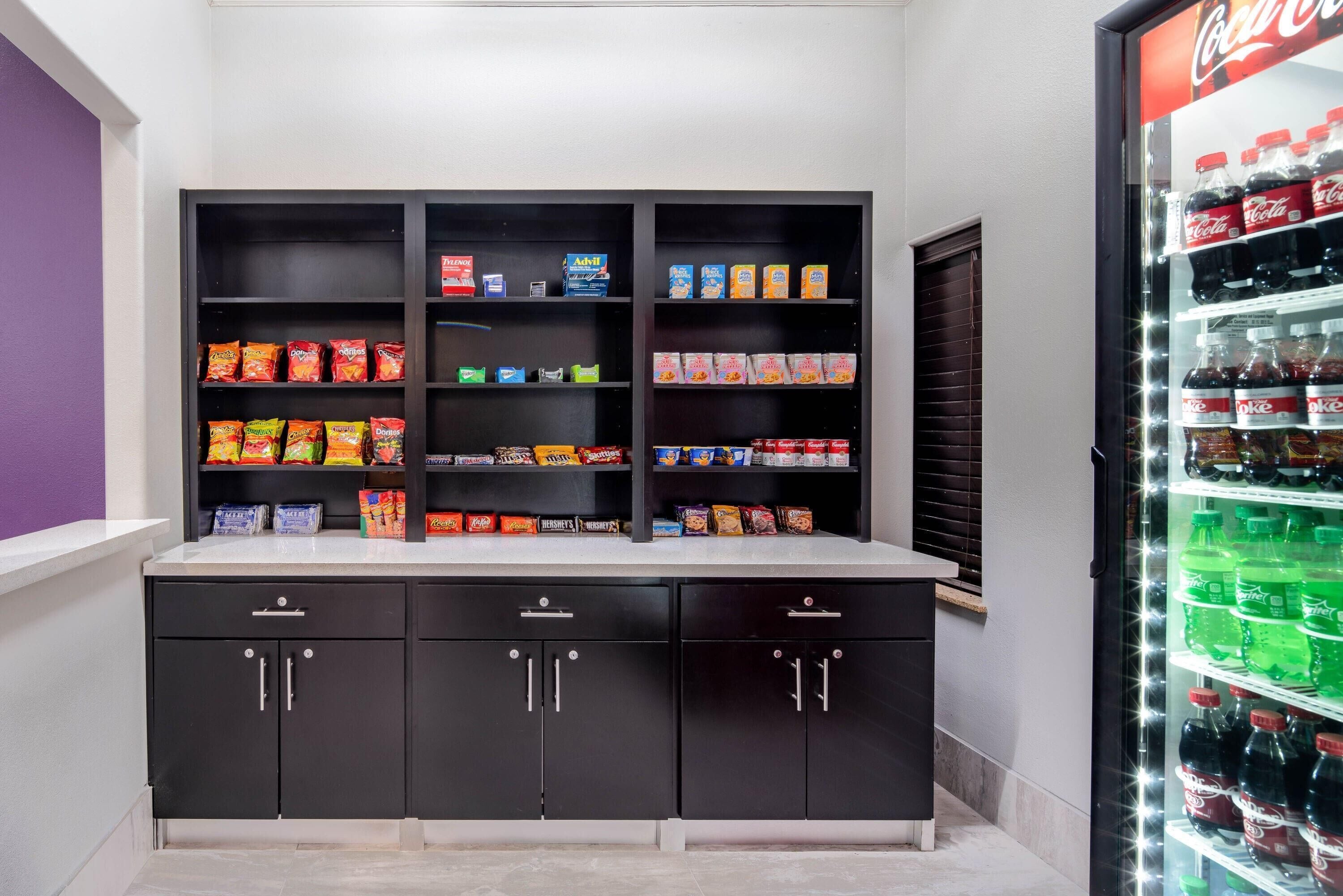 A kitchenette with a refrigerator stocked with beverages, a shelf with snacks, and a window with blinds.