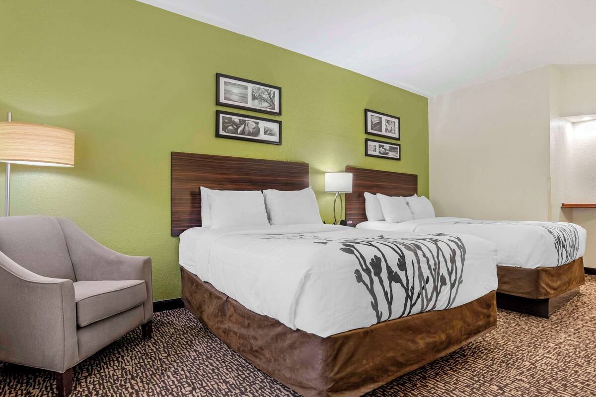 A hotel room with two beds, a chair, and a lamp. There are framed pictures on the wall.