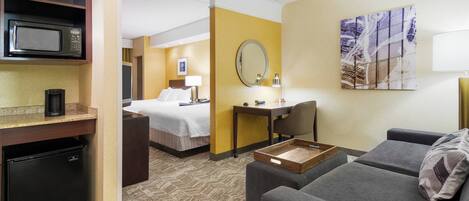 A hotel room with a kitchenette, a bed, a desk, a chair, a sofa, and a painting on the wall.