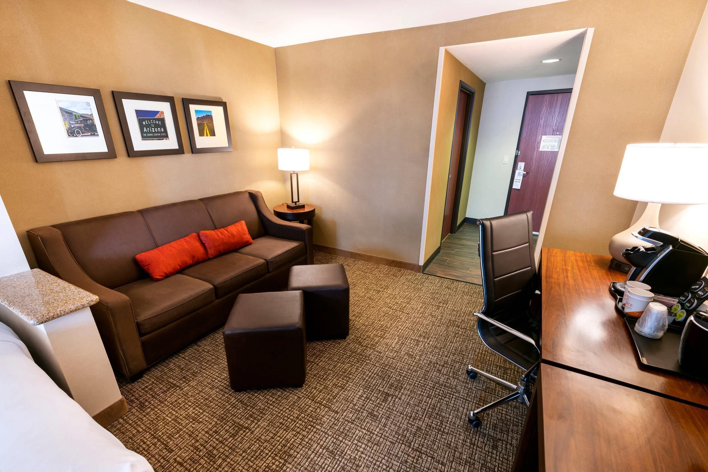 Habitación de hotel con sofá marrón, una mesita, un escritorio con lámpara y cuadros en la pared.