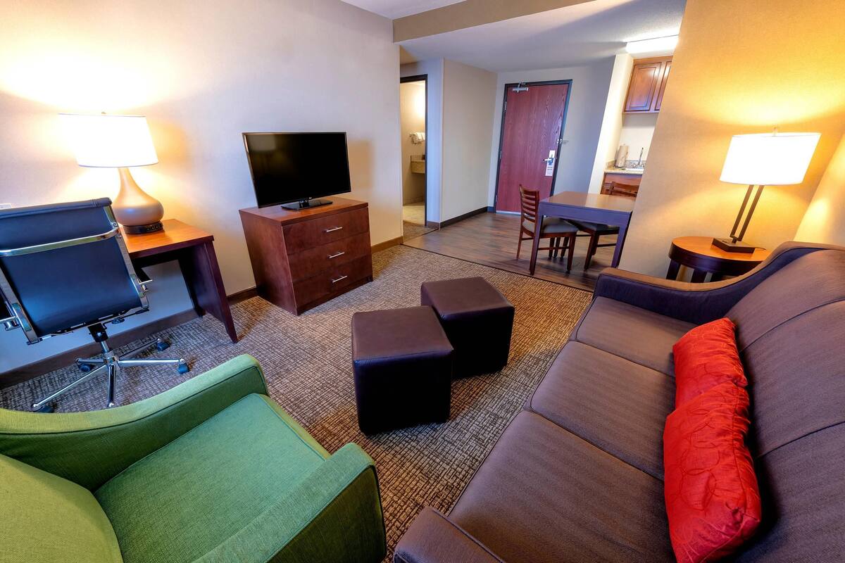 Ein Hotelzimmer mit einem grünen Sessel, einem braunen Sofa mit einem roten Kissen, einem Fernseher auf einem Holzboden, einem Esstisch mit Stühlen und einem kleinen Tisch mit einer Lampe.