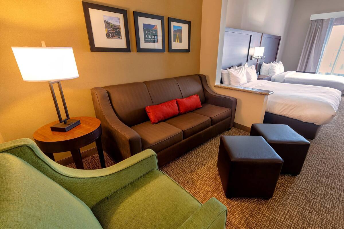 Ein Hotelzimmer mit braunem Sofa, grünem Sessel, zwei schwarzen Hocker, einem kleinen runden Tisch mit Lampe und einem Bett mit weißer Bettwäsche.