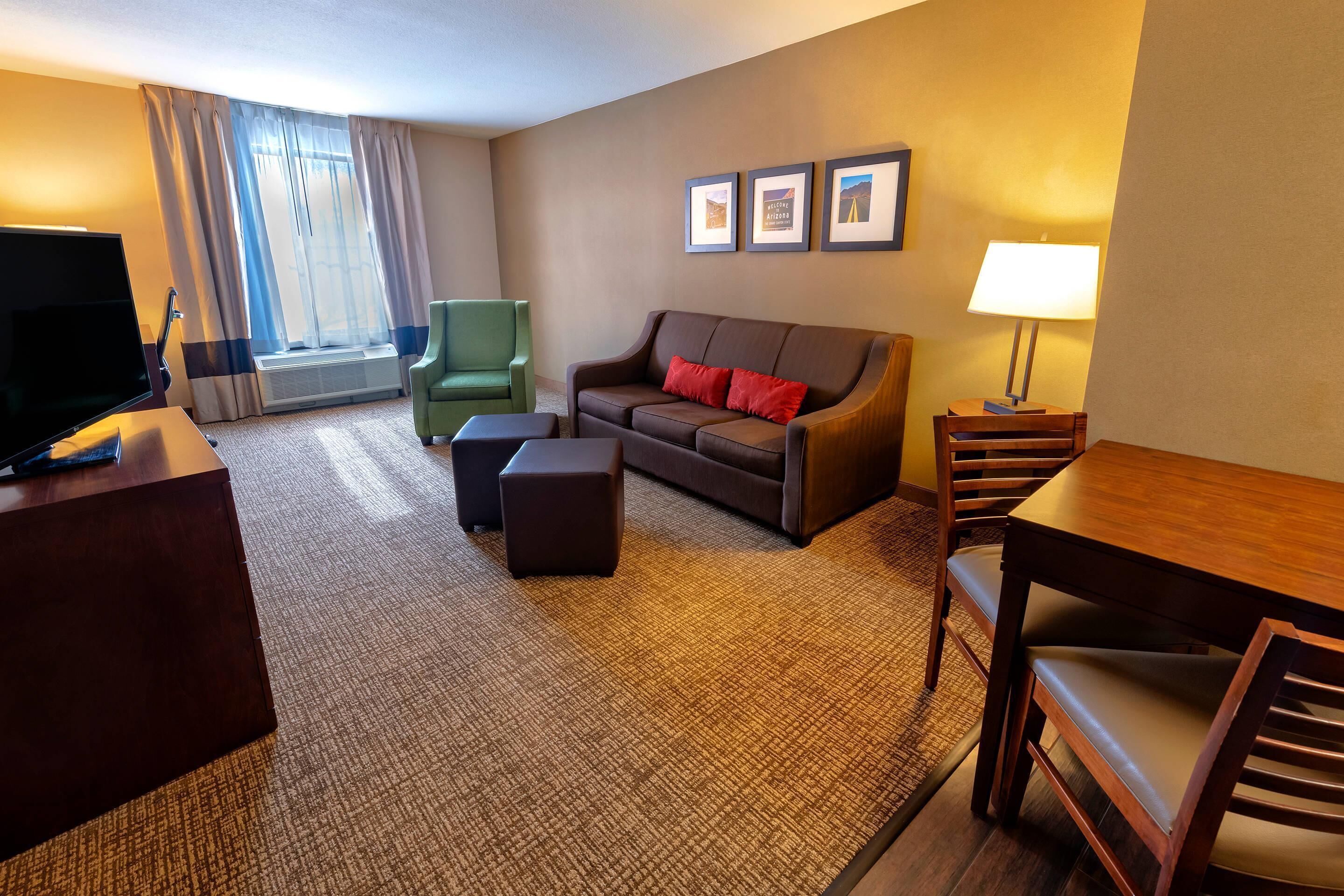 A hotel room with a flat-screen TV, a sofa with red cushions, a green armchair, a wooden dining table, and a lamp.
