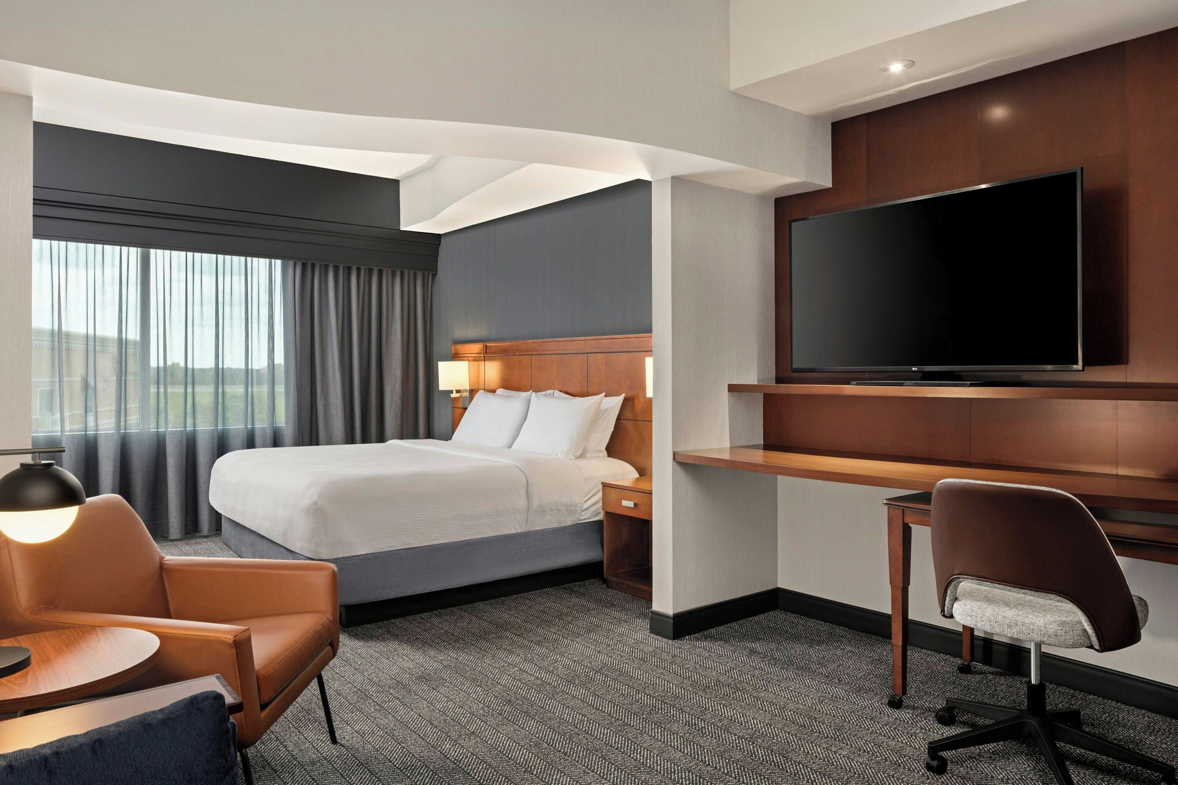 A hotel room with a bed, a desk with a TV, a chair, and a lamp.