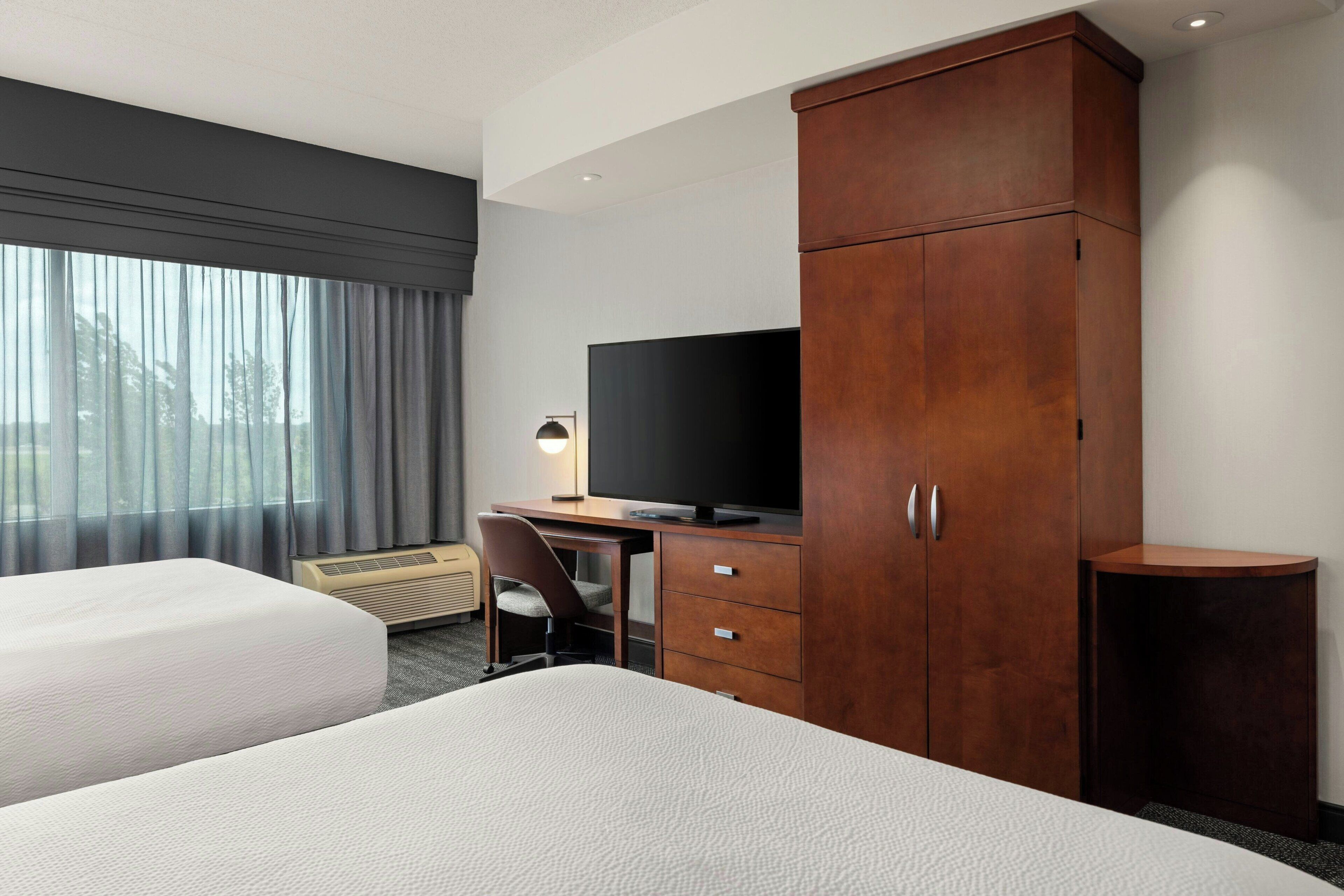 Habitación de hotel con dos camas, un escritorio con televisor, un armario y una ventana con cortinas.