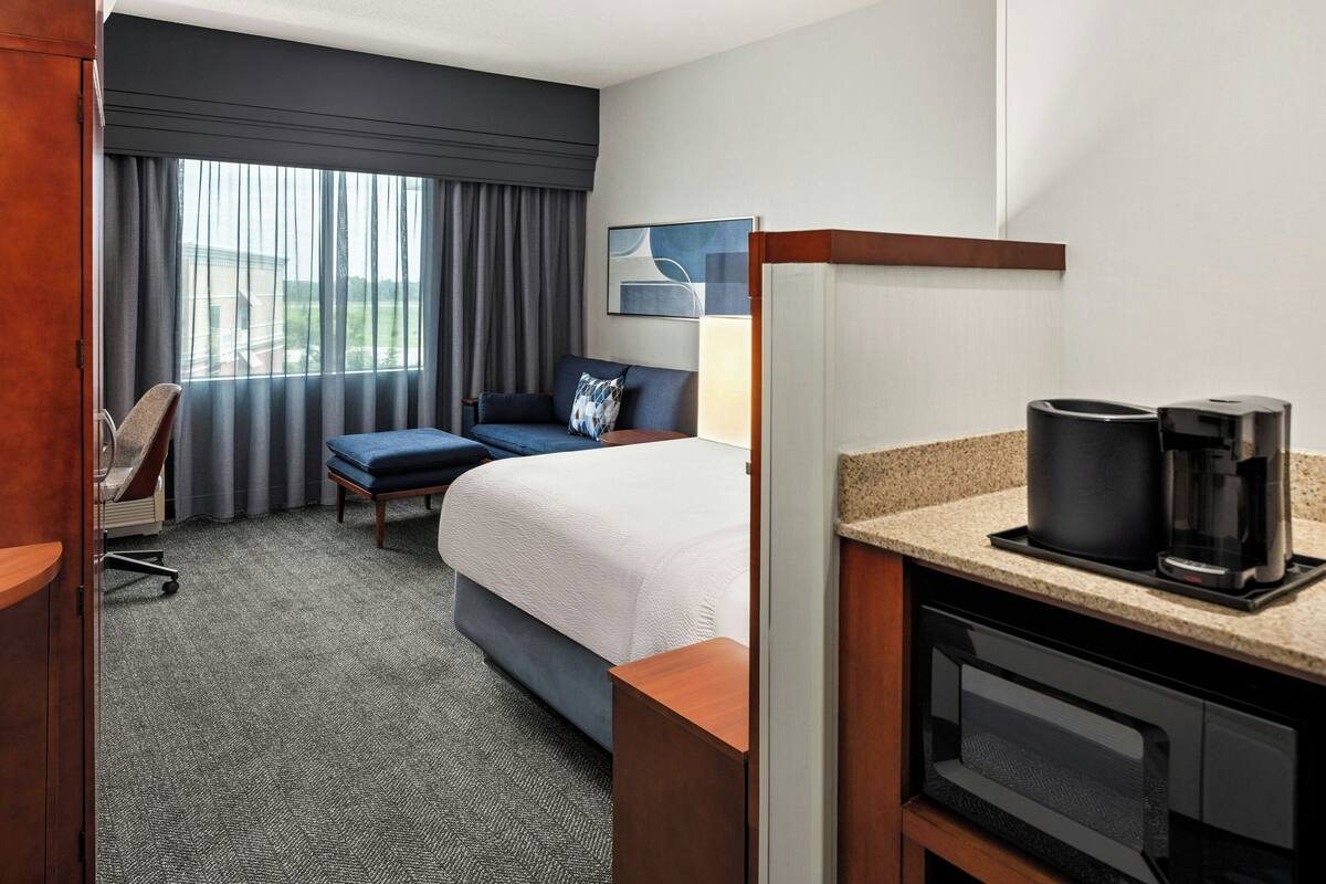 Een hotelkamer met een bed, een bureau met koffiezetapparaat, een stoel, een bureaulamp en uitzicht buiten het raam.