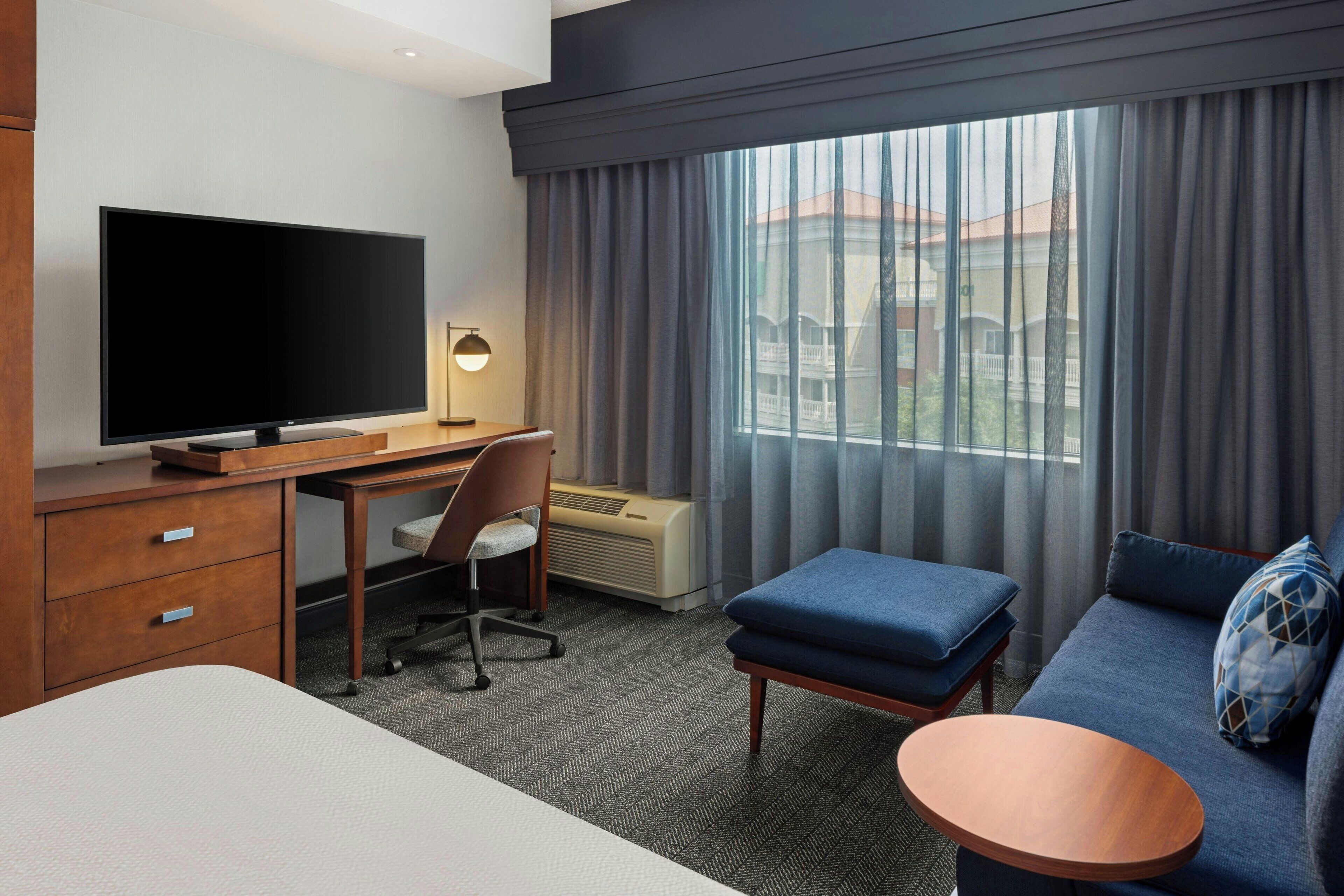 Habitación de hotel con una televisión de pantalla plana grande, un escritorio, una silla, un sofá, una mesita redonda y una ventana con cortinas.