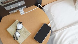 A neatly made bed with white sheets, a bedside table with a smartphone and glasses, and a power outlet with a charging cable. - Comfort Hotel Tokyo Kanda (Tokyo)
