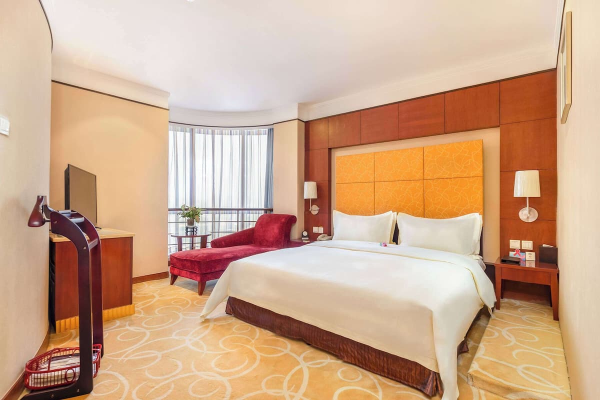 A hotel room with a large bed, a red armchair, a desk, and a window with curtains.