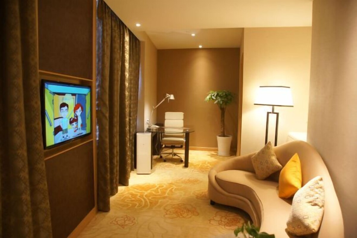 A hotel room with a television, desk, chair, lamp, potted plant, and a sofa with cushions.