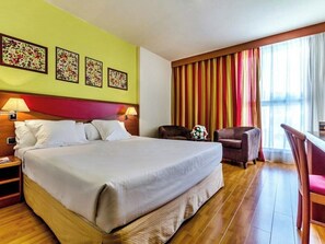 Ein Hotelzimmer mit einem großen Bett, zwei Nachttischen, einem Schreibtisch mit Stuhl, einem Fenster mit Vorhängen und floraler Wandgestaltung.