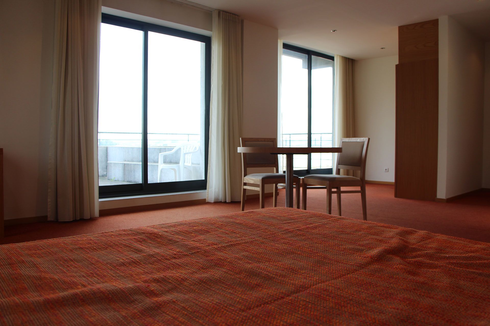 Una habitación de hotel con una cama grande, dos sillas, una mesita y ventanas amplias con cortinas.