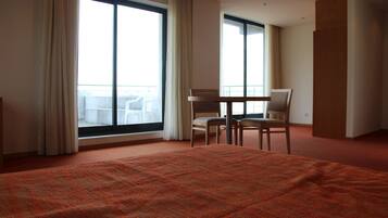 Una habitación de hotel con una cama grande, dos sillas, una mesita y ventanas amplias con cortinas.