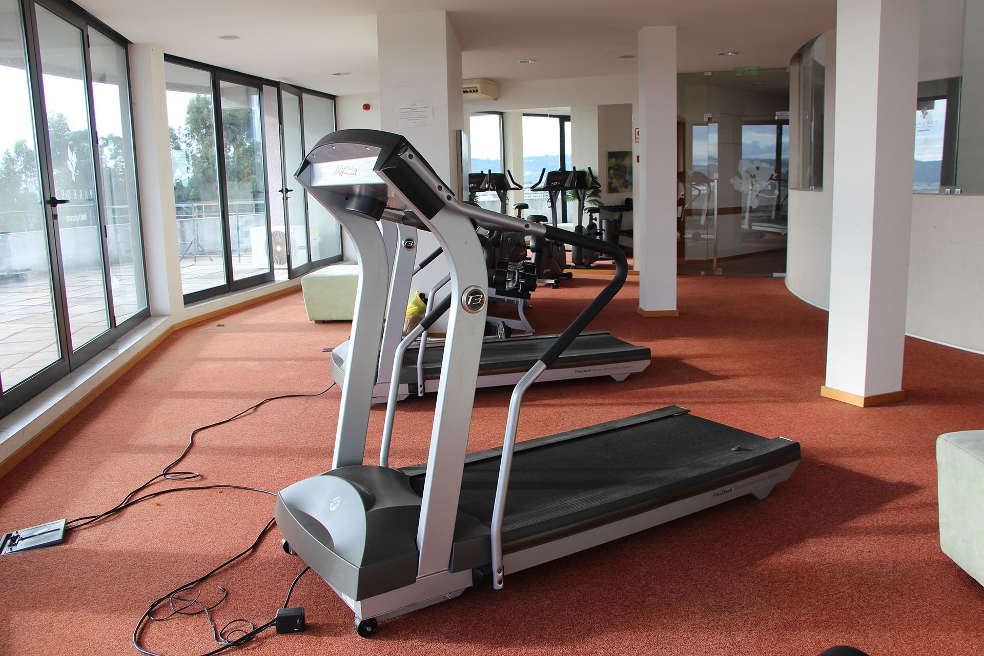 Ein Fitnessstudio mit Laufbändern und Trainingsgeräten, großen Fenstern und einem Teppichboden.