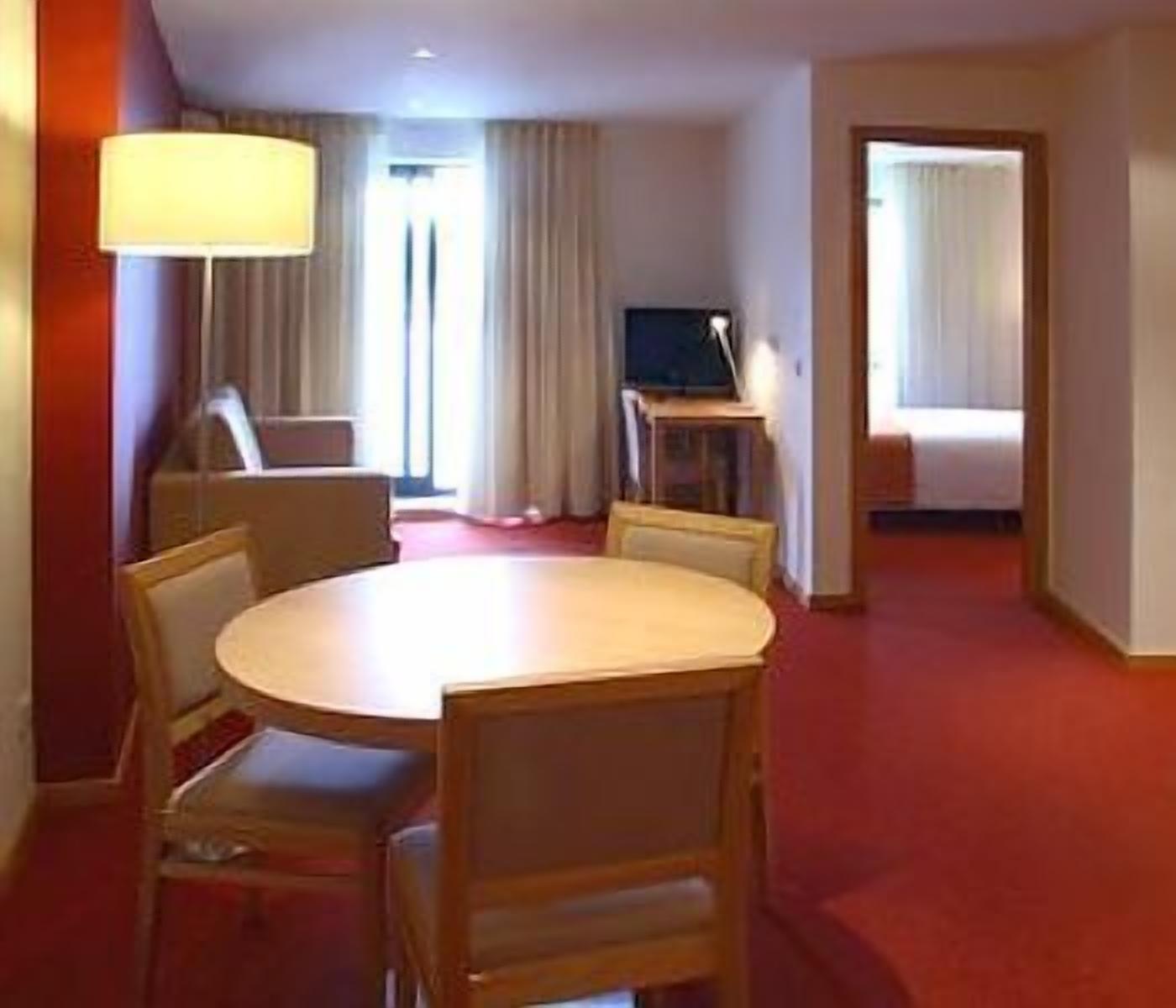 A hotel room with a round dining table, chairs, a TV, and a bed.
