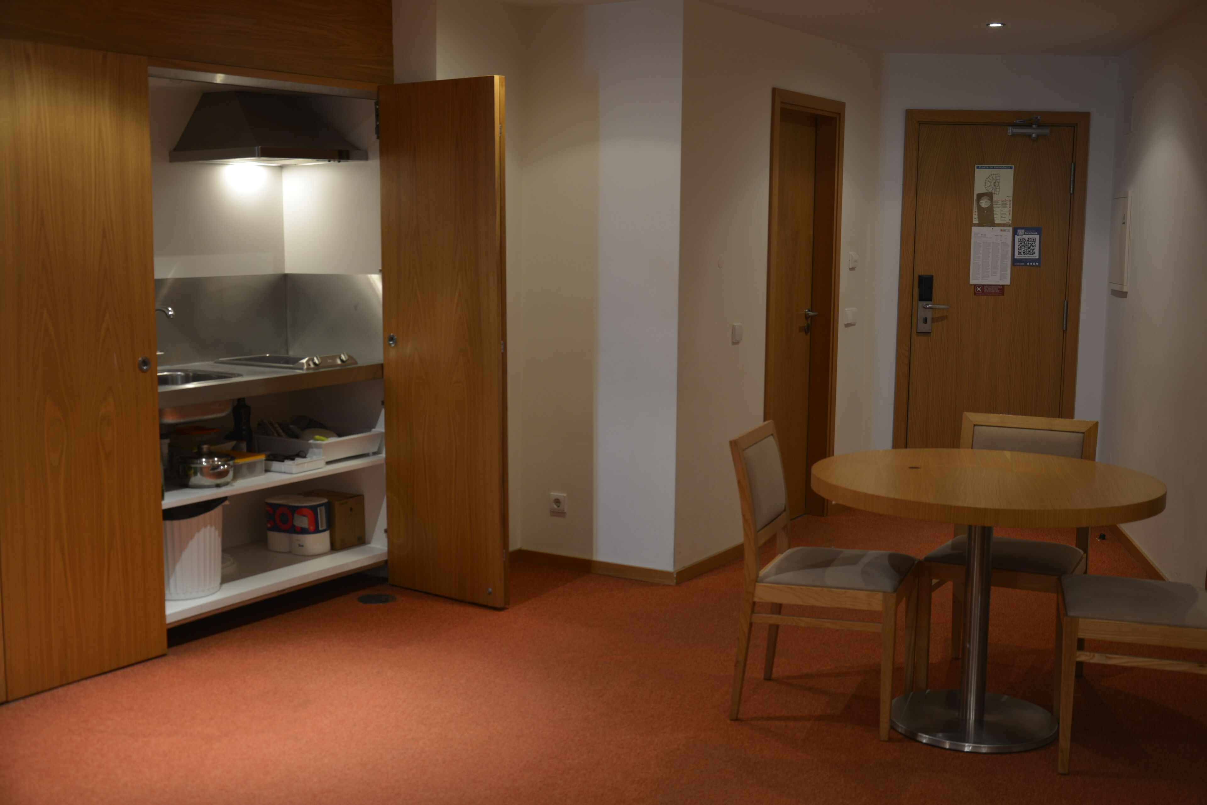 A room with a wooden cabinet, a round table with chairs, and a door with a QR code.